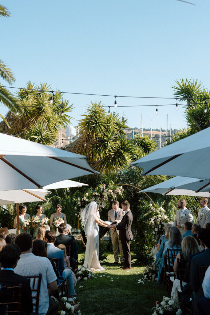 Bride and groom exchanging vows during an outdoor ceremony at Glen Cove Marina, surrounded by palm trees and sailboats in Northern California