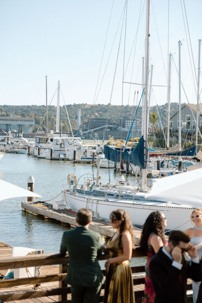Couple arriving by sailboat to cocktail hour at Glen Cove Marina as guests wave from the shoreline during golden hour