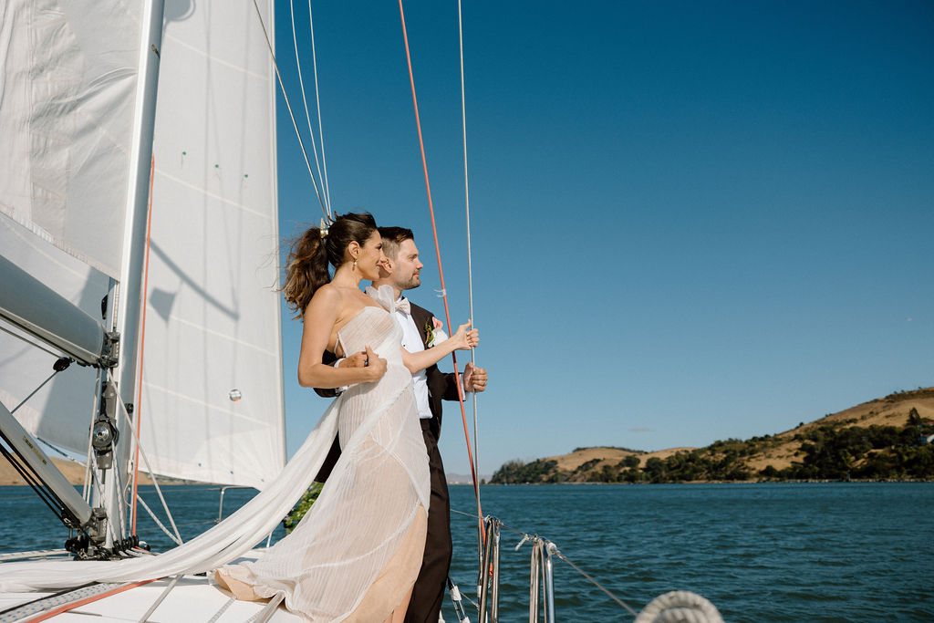 Bride and groom sailing together on San Francisco Bay after their wedding ceremony, enjoying a just-married cruise. Bride wearing a flowing wedding gown in ocean breeze during a Northern California sailboat wedding