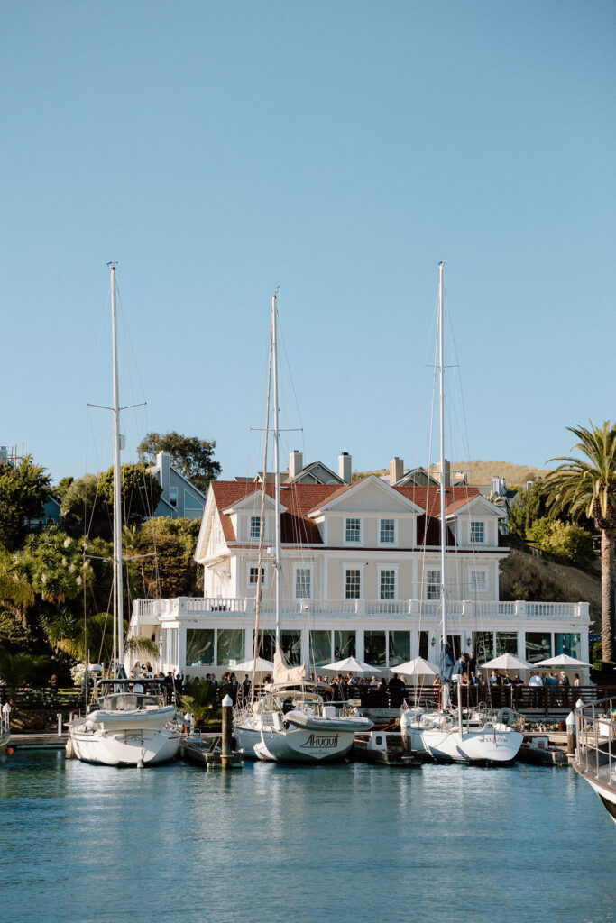 Couple arriving by sailboat to cocktail hour at Glen Cove Marina as guests wave from the shoreline during golden hour