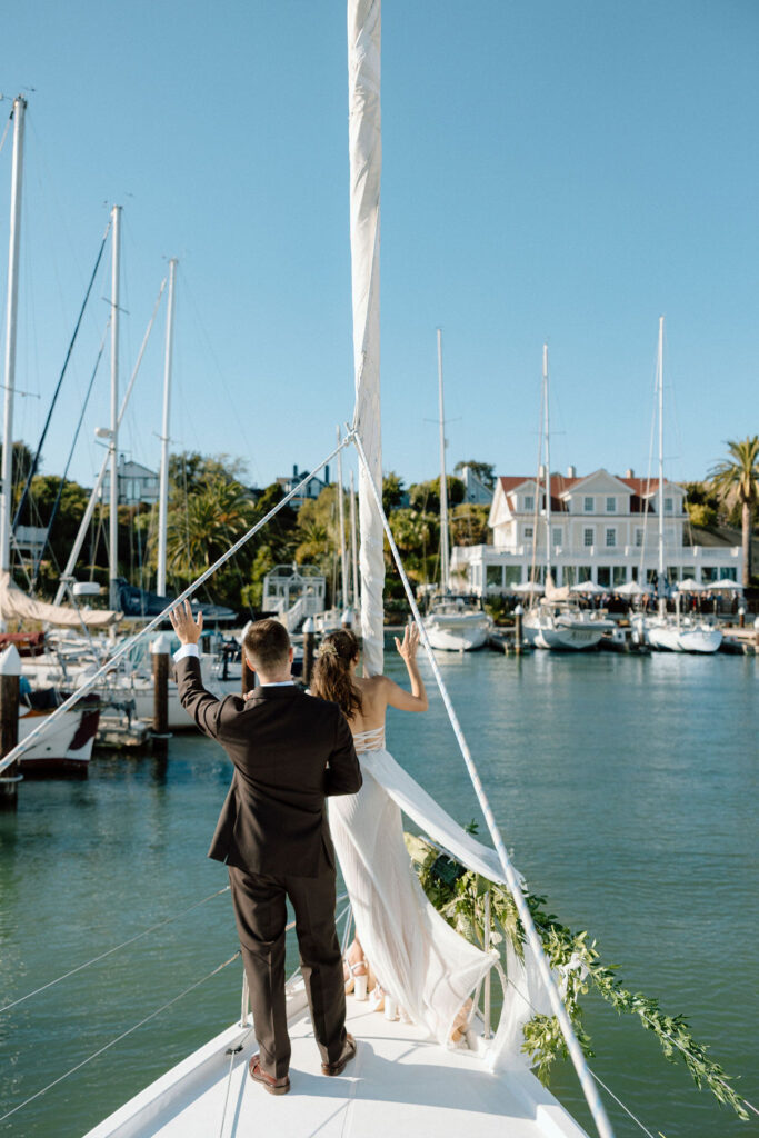 Couple arriving by sailboat to cocktail hour at Glen Cove Marina as guests wave from the shoreline during golden hour