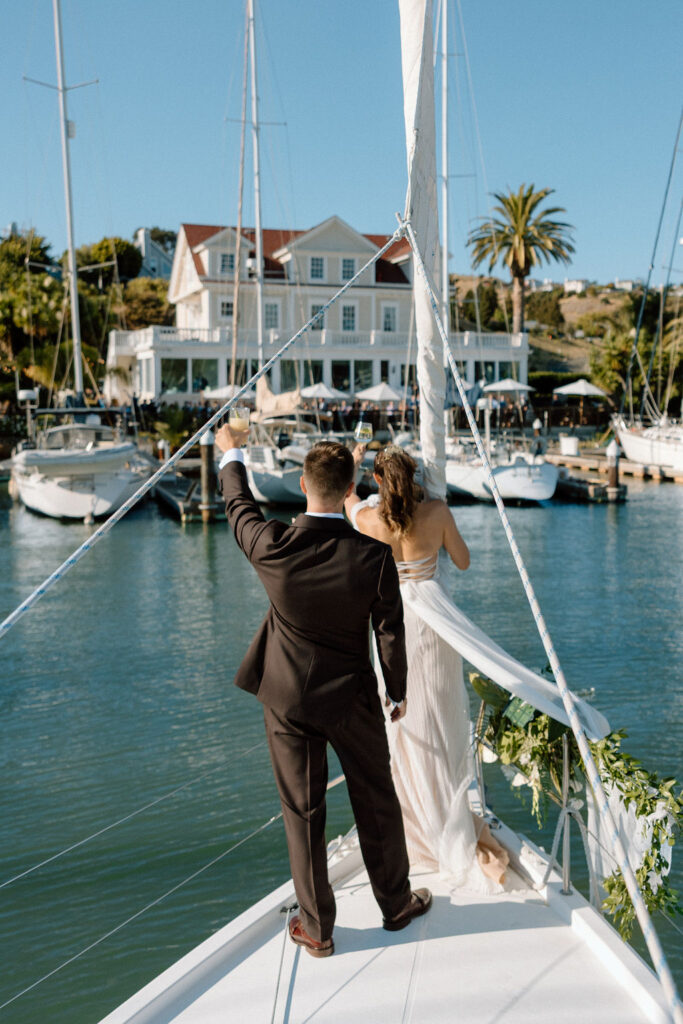 Couple arriving by sailboat to cocktail hour at Glen Cove Marina as guests wave from the shoreline during golden hour