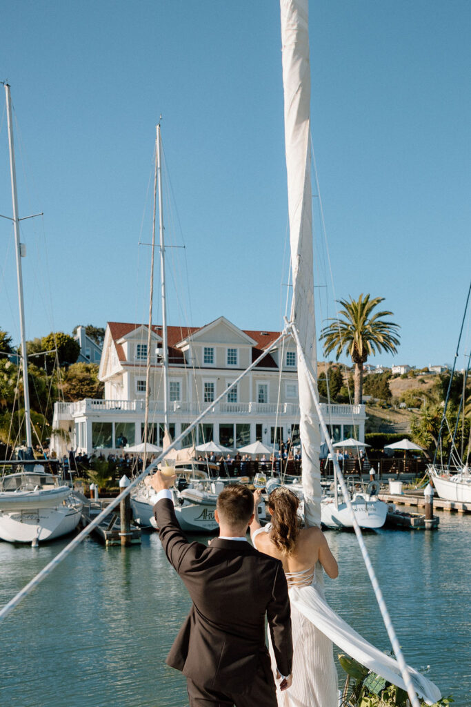 Couple arriving by sailboat to cocktail hour at Glen Cove Marina as guests wave from the shoreline during golden hour