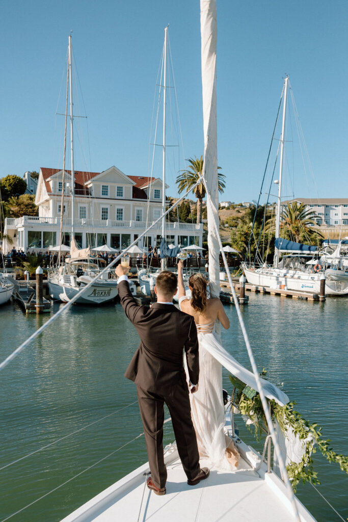 Couple arriving by sailboat to cocktail hour at Glen Cove Marina as guests wave from the shoreline during golden hour
