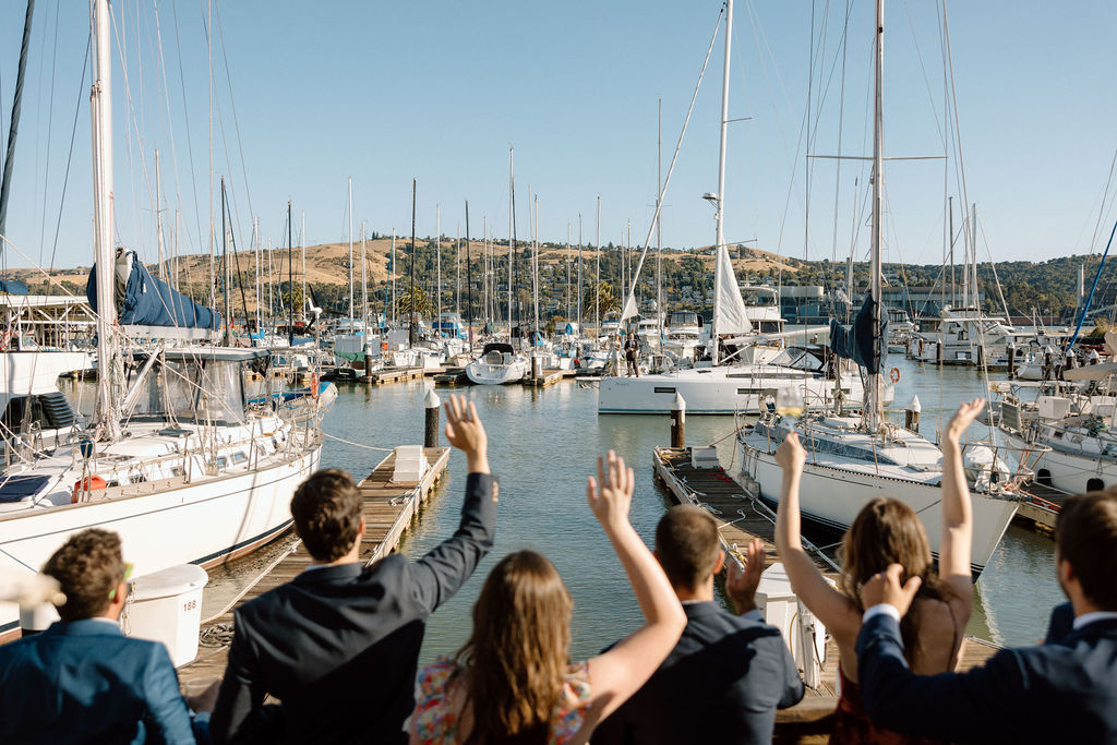Couple arriving by sailboat to cocktail hour at Glen Cove Marina as guests wave from the shoreline during golden hour