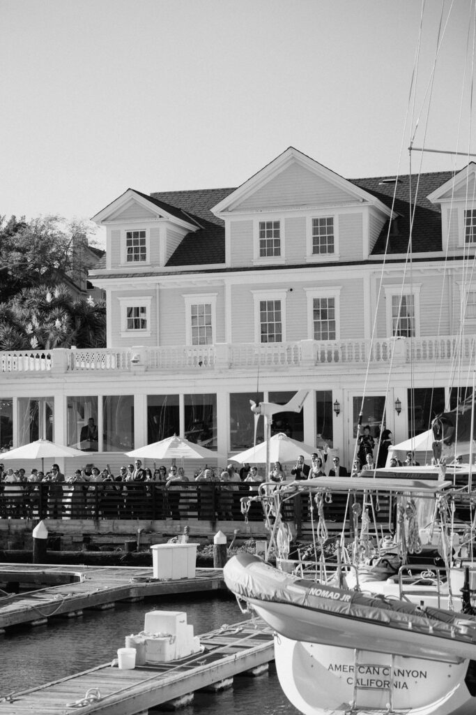Couple arriving by sailboat to cocktail hour at Glen Cove Marina as guests wave from the shoreline during golden hour