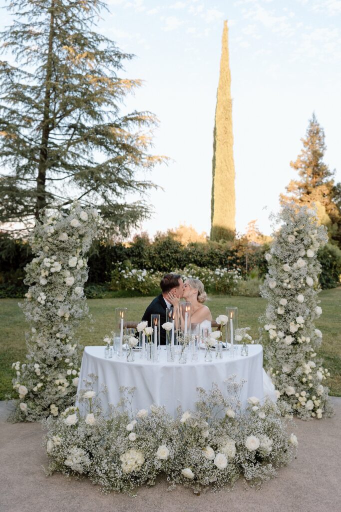 bride and groom kiss at reception table with florals and candles surrounding them 