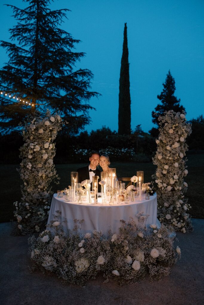 bride and groom smile at reception table with florals and the candles lighting them up at blue hour