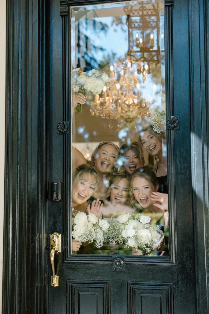 Bridesmaids look through the window smiling and showing their florals as bride does first look with her dad on her wedding day