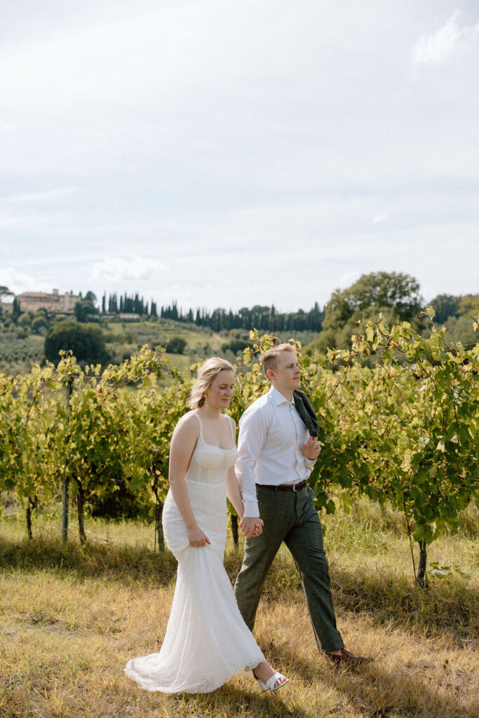 Bride and groom walking hand in hand through sunlit vineyards in Tuscany during an intimate Italian destination wedding.