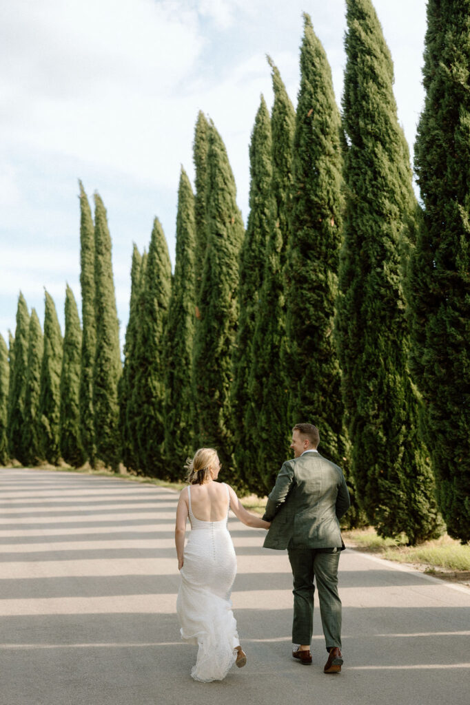Bride and groom walking hand in hand through cypress-lined Tuscan landscape moments after saying vows at their destination elopement